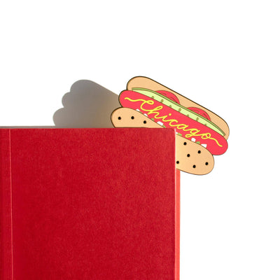 A red book with the Pineapple Sundays Design Studio Chicago Style Hot Dog Enamel Brass Bookmark peeking out from the top right corner, set against a white background.