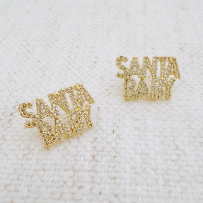 The Ellison+Young Santa Baby Crystal Stud Earrings feature gold-tone letters adorned with sparkling rhinestones, perfect for festive holiday wear, showcased on a textured white fabric background.