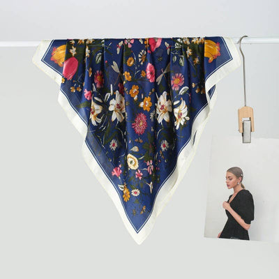 The LARGE FLORAL PRINT BANDANA SCARF by JOSSLYN by wall to wall, featuring a blue background and colorful floral design, hangs over a white rod beside a clipped photo of a woman in a black dress.