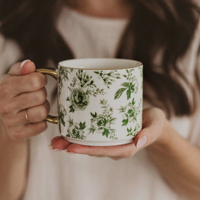 A person is holding a Sweet Water Decor Green Delft Coffee Mug featuring a gold handle, while wearing a light-colored shirt. Their brown hair is loosely styled, hands neatly manicured. The softly blurred background enhances the elegance of the fine bone china mug in their grip.