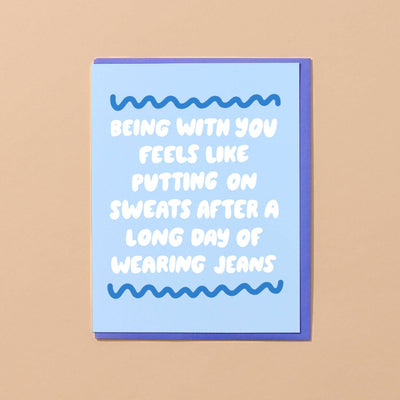 Designed by Kiwi Schloffel, the Putting On Sweats Love Card from And Here We Are features a light blue front with the message Being with you feels like putting on sweatpants after a long day of wearing jeans in white letters, set against blue wavy lines and a cozy beige background.