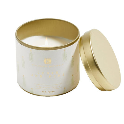 A 9oz Cedar & Pine Candle Tin by Hillhouse Naturals/Field+Fleur glows in a gold container with a white label featuring green tree illustrations and gold text. The lid is set aside. Proudly made in the USA.