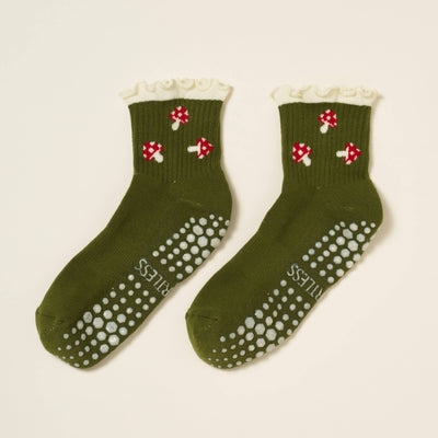The Effortless Club Mushroom Ruffle Grip Sock is a green organic cotton pair with white ruffled cuffs, red and white mushroom designs, cushioned footbed, grippy dots, and "CARELESS" printed on the soles.
