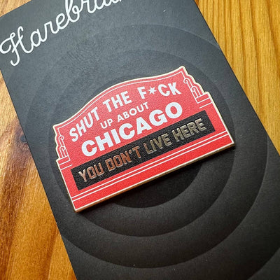 The Harebrained! STFU About Chicago Northside Pin, a red and black sign-shaped magnet designed in Chicago, sits on a wooden surface. It reads “Shut the f*ck up about Chicago, you don’t live here,” with censored text.