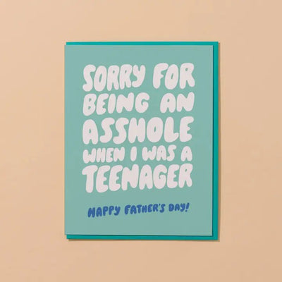 The Asshole Teenager Fathers Day Greeting Card by And Here We Are is letterpress printed on premium cotton paper with a teal background and bold white text reading, Sorry for being an asshole when I was a teenager, followed by smaller blue text saying, Happy Fathers Day! The card is A2-sized and humorously toned.