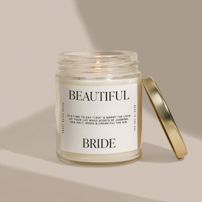 The Sweet Water Decor Beautiful Bride 9 oz Soy Candle features a gold-lidded glass jar. Infused with premium fragrance oils, it emits jasmine, sea salt, wood, and cream scents, ideal for spa-like relaxation against a neutral beige backdrop.