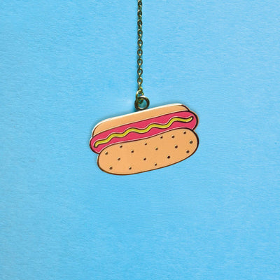 The Chicago Style Hot Dog Bookmark Charm by Pineapple Sundays Design Studio is a hard enamel metal charm shaped like a hot dog with mustard and ketchup, shown hanging against a light blue background.