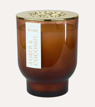 The 21 oz Amber & Coconut Maeve Glass Candle by Elum, crafted with soy wax and Aura Pure technology for a clean burn, is housed in a brown glass jar with a gold lid. Its label elegantly shows the name in white text.