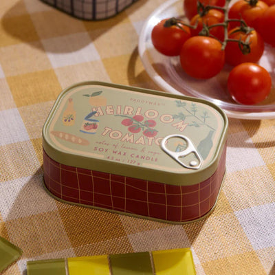 A Paddywax Heirloom Tomato Bistro Printed Tin Candle rests on a yellow checkered tablecloth near cherry tomatoes, filling the air with a fresh tomato scent. The candle comes in a tin with vintage graphics and a pull-tab lid.