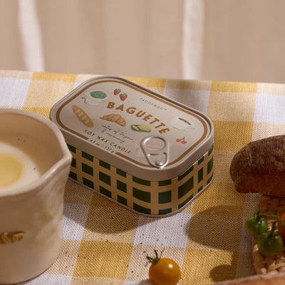 A Paddywax French Baguette Bistro Printed Tin Candle sits on a yellow and white checkered tablecloth next to a candle, a baguette piece, and a small yellow tomato.