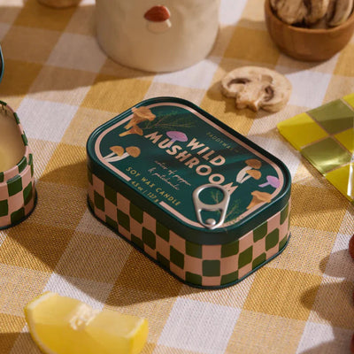 A Paddywax Wild Mushroom Bistro Printed Tin Candle rests on a yellow checkered cloth, surrounded by a lemon wedge, mushrooms, and a ceramic container, inviting you to experience its unique wild mushroom scent.