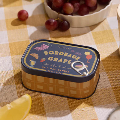 The Paddywax Bordeaux Grape Bistro Printed Tin Candle sits on a yellow checkered tablecloth, surrounded by grapes, a lemon wedge, and fresh fruit in soft light—ideal for fans of fragrant tin candles.