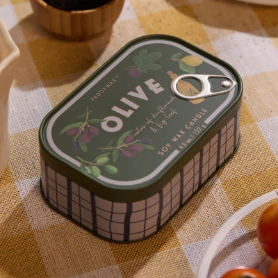 A Fig And Olive Bistro Printed Tin Candle by Paddywax rests on a yellow and white checkered tablecloth, surrounded by plates, with olive illustrations decorating its lid.
