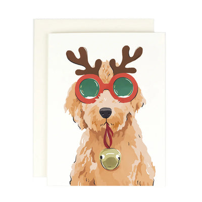 The Reindeer Doodle Greeting Card by Amy Heitman features a fluffy tan dog with reindeer antlers, festive green sunglasses, and a gold bell on a white background, perfect for sending with its blank interior and matching envelope.