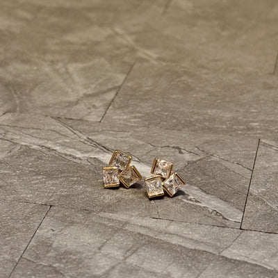 Golden Stellas baguette cluster earrings feature square-cut, gold-framed brass with clear gemstones, displayed on a gray stone-textured surface.