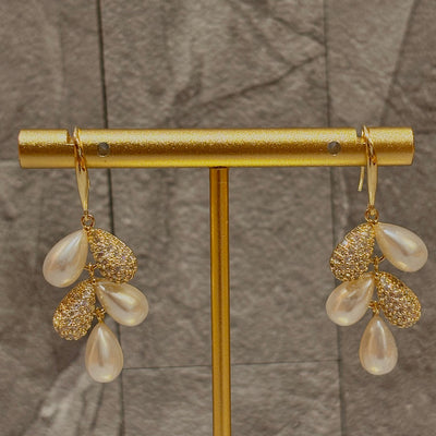 The Vera Pearl Cluster Earrings by Golden Stella feature three teardrop faux pearls and two gold, gem-studded accents on each 1.5 earring, shown on a gold stand with a textured gray backdrop.
