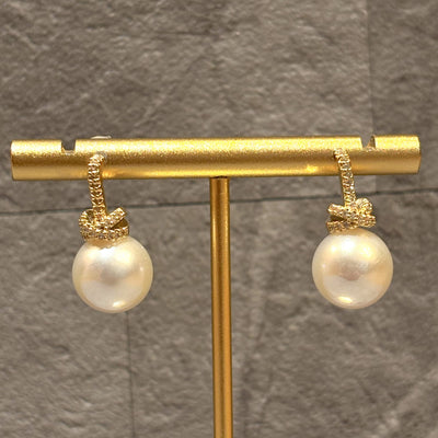 Lora Pearl Earrings by Golden Stella feature large round white pearls on a gold-dipped brass base, elegantly displayed on a gold stand against a textured gray background.