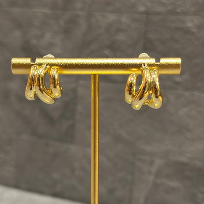 The Bella Huggie Earrings by Golden Stella feature a chunky, twisted design in gold-dipped brass with Silver 925 posts, shown on a gold stand against a textured gray background.