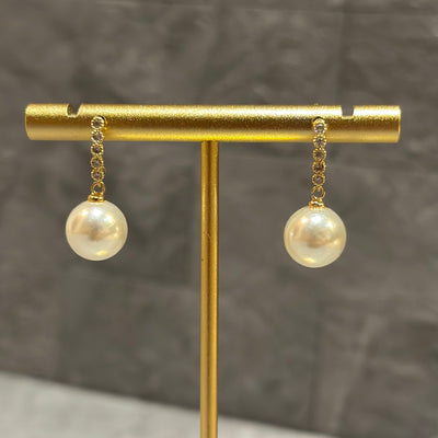 The Danica Pearl Earrings by Golden Stella feature gold chain links, large round pearl drops, and a brass base. Each earring measures 3/4 long and is shown on a gold stand against a textured gray background.