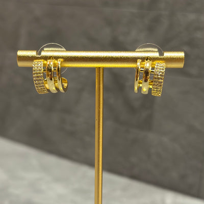 The Trifecta Huggies by Golden Stella are gold hoop earrings featuring multiple rows with small clear stones, crafted from brass dipped in gold, and shown on a gold stand against a neutral gray background.