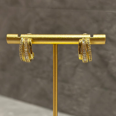 The Trianon Earrings by Golden Stella are gold hoops featuring rows of rectangular and round clear stones on a brass base, showcased on a gold stand with a gray textured background.
