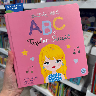 A hand holds the ABCs of Taylor Swift Book Board Book by Ingram Publishing, showing an illustrated blonde girl, musical notes, and stars on the cover. Bookshelves appear in the background.