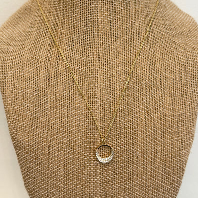 The Eclipse Necklace by Golden Stella features a gold-tone adjustable chain and circular pendant partly set with clear stones, crafted from brass and displayed on a textured burlap jewelry bust.