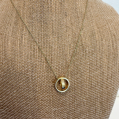 The Golden Stella nested rings necklace features two interlocking rings—one polished, one with sparkling stones—on a delicate 16-inch gold chain. Crafted with a brass metal base and shown on a burlap bust, it adds an elegant touch to any look.