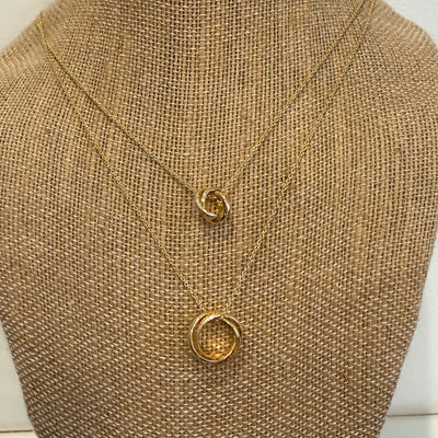 The Golden Stella layered interlocking rings necklace features two gold chain necklaces with an adjustable length for layered styling. Each has a brass base and circular, intertwined pendants, showcased on a textured burlap stand.