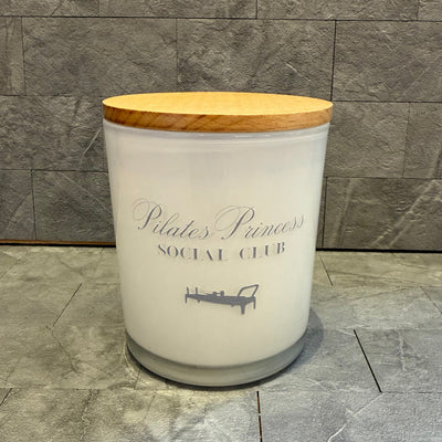The Unplug Soy Candles Pilates Princess Social Club Soy Candle, featuring a wooden lid and label with a Pilates reformer illustration, is displayed on gray tiles—ideal for your self-care routine.