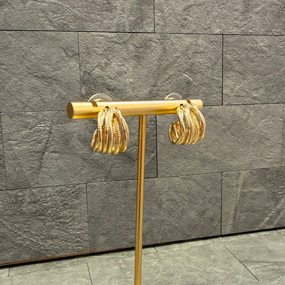 The Golden Stella Textured Layer Hoop Earrings, crafted from zinc with a twisted design and 0.75 diameter, are displayed on a sleek gold stand against a textured gray stone background.