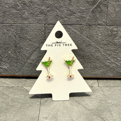 A pair of Christmas Martini Enamel Stud Earrings by Prep Obsessed Wholesale, featuring gold and green enamel with a red accent, are showcased on a white tree-shaped card reading "The Fig Tree" against a gray textured background.