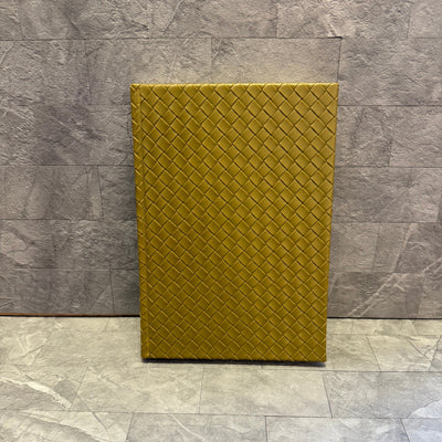 The Punch Studio Woven Vegan Leather Journal in olive, featuring a textured diamond pattern, stands upright against a gray stone-tiled wall and floor.