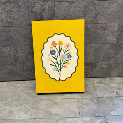 The Softcover Journal - Flowers by Punch Studio features a yellow cover with colorful flowers in a scalloped oval frame, shown upright against a textured gray background.