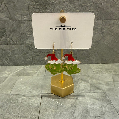 A pair of 1.5-inch green Grinch face earrings with red Santa hats from Golden Stella hang on a gold stand, displayed on a card reading "The Fig Tree" against a gray tile background. Product: The Grinch Earrings.