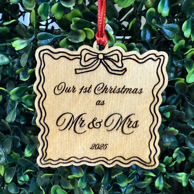 The Jim Harris "Our First Christmas as Mr & Mrs" Ornament features a scalloped wooden design with bow, gold-edge satin ribbon, and “Our 1st Christmas as Mr & Mrs 2025” text. Measures 2.75 x 2.75 inches. Green foliage background.