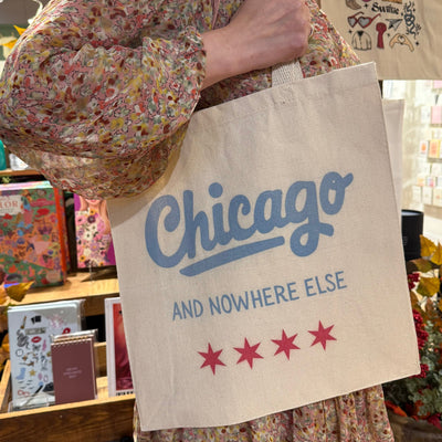 A person in a floral dress holds the Chicago and Nowhere Else Tote Bag by Lucky Luna Apparel, featuring four red stars. Colorful books and decorative items fill the shelves in the background.