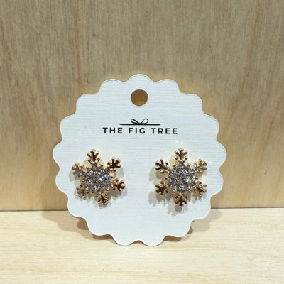A pair of sparkling snowflake earrings by Golden Stella in gold and silver with stones, displayed on a white scalloped card, set against a light wooden background.
