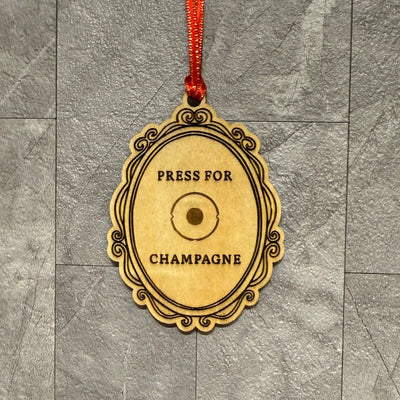 The Jim Harris Press for Champagne Ornament is a 3" x 2.25" laser-engraved birchwood plaque with a central button and "PRESS FOR CHAMPAGNE" text, hanging from a red ribbon against gray tiles.