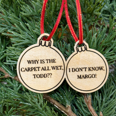 Two "todd & margo ornament" birch pieces by Jim Harris, laser-engraved with “WHY IS THE CARPET ALL WET, TODD??” and “I DONT KNOW, MARGO!”, hang from red ribbons on a green pine tree.