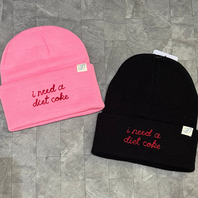 Two I Need a Diet Coke Chainstitch Embroidered Beanies by Daily Disco—one pink, one black—rest on a gray textured surface. Each features “i need a diet coke” in red cursive and a small white cuff tag with the D logo.