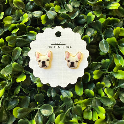French Bulldog Earrings by BETTYOH INC are displayed on a round card, surrounded by artificial green leaves. These hypoallergenic earrings feature French bulldog faces and titanium posts for comfortable wear.