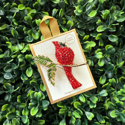 The Golden Stella cardinal brooch is a 2-inch, gold-plated pin featuring a red cardinal with green leaves. It comes attached to a cream card, tied with a gold ribbon, and displayed against green artificial foliage.