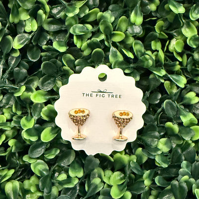 Espresso martini-shaped earrings by Golden Stella are displayed on a round white card, set against a vibrant green leafy background.