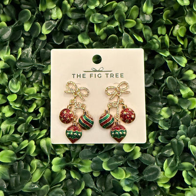 Golden Stella’s triple ornament Christmas earrings feature bow-shaped tops, colorful charms, and hypoallergenic titanium posts. Displayed on a white card, they’re set against green leaves for a festive touch.