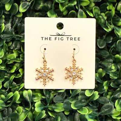 Golden Stella Gold Snowflake Earrings feature small center crystal rhinestones and hypoallergenic brass hooks, displayed on a card amid green leafy plants.