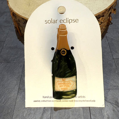 The Solar Eclipse Hand-painted Champagne Claw Hair Clip is made from biodegradable acetate and recycled wood pulp, shaped like a champagne bottle with the label “Champagne Brut 1926 France.”.