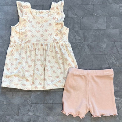 The Angel Dear Vintage Mini Bows Square Neck Ruffle Tank, showcasing a pink bow pattern and frilled sleeves, pairs beautifully with rib modal pink bike shorts featuring wavy hems. This charming, machine-washable outfit is displayed on a textured gray surface.