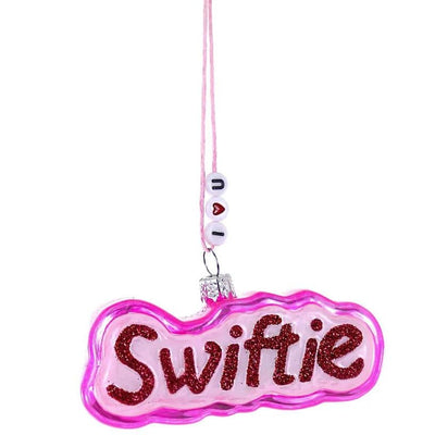 The Cody Foster Pink Swiftie Ornament is a 3.5-inch glass decoration in pink and white, featuring "Swiftie" in red glitter and hanging from a beaded string that spells I ♥ U.