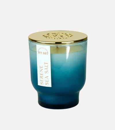 Elums Serene Sea Salt 7 oz Maeve Glass Candle features a blue ombre design with a gold lid. A tag labeled SERENE SEA SALT № 007 highlights its Aura Pure technology, offering a calming experience.